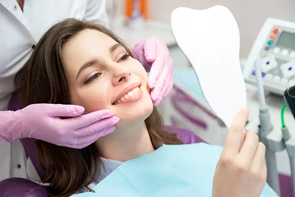 A smiling woman holding a tooth-shaped mirror while a dentist adjusts her face, showcasing her dental results in a clinic.
