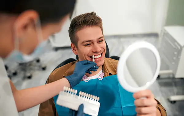 Male patient holding a mirror and smiling as a dentist matches a dental veneer shade to his teeth using a shade guide.